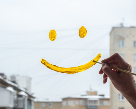 A womans hand draws a smiling face with yellow paint on the glass window with a view of city houses, close-up.の写真素材