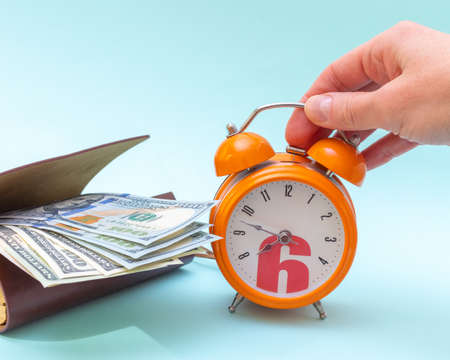 Time management concept. A hand holds an orange alarm clock and dollars in an open wallet on a blue backgroundの写真素材