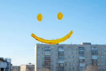 A smiling face painted in yellow paint on a glass window with a view of city houses, close-up. A symbol of hope, happiness, joy.の写真素材