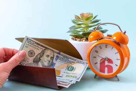 Time management concept. Hand takes money from an open wallet, orange alarm clock and succulent plant on a blue background, close-upの写真素材