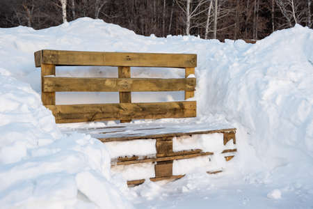 Wooden bench from pallets in a snowdrift after a snowfall in a park in the forest. Snowfall and drifts symbolの写真素材