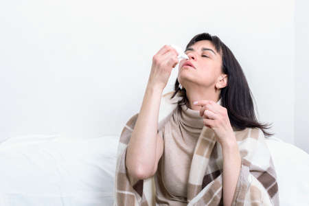 A sick woman covered with a blanket sprinkles a medicinal solution on her nose. A Caucasian woman has a high fever, runny nose and common cold. Seasonal illness, allergy, flu conceptの写真素材
