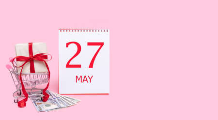 27th day of may. A gift box in a shopping trolley, dollars and a calendar with the date of 27 may on a pink background. Spring month, day of the year concept.の写真素材