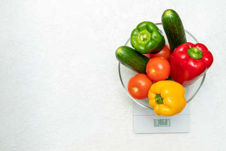 Multicolored fresh sweet peppers, tomatoes, cucumbers in a transparent bowl on a kitchen scale on a whiteの写真素材