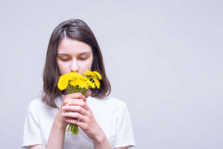Pretty brunette girl holding yellow wildflowers of dandelions and inhaling their aroma on a light backgroundの写真素材