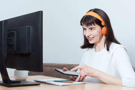 A smiling woman in orange headphones sitting in front of the monitor and holding a smartphone for...