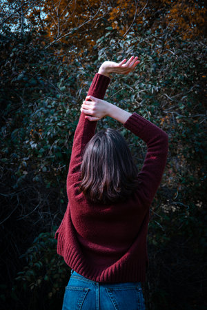 Happy young brunette girl in a brown sweater standing with raising her hands up in a forest scenery....