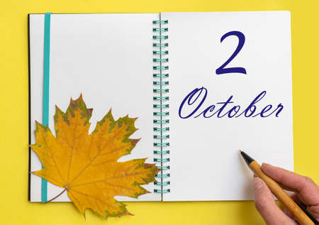2nd day of october. Hand writing the date 2 october in an open notebook with a beautiful natural maple leaf on a yellow background. Autumn month, day of the year concept.の写真素材