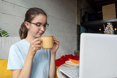 Beautiful girl student drinking coffee, tea, cocoa from a mug while sitting in front of a laptop. Business concept.の写真素材