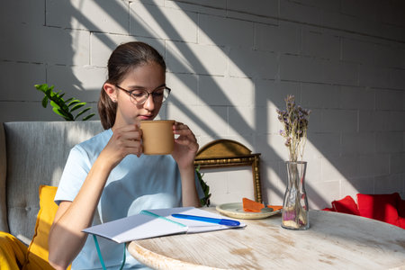 Beautiful girl student drinking coffee and writing a task in a notebook while sitting in a cafe with...