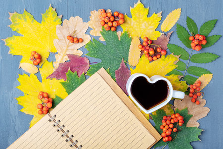 White mug in the shape of a heart with coffee or tea and a blank notepad on the background of autumn dry leavesの写真素材