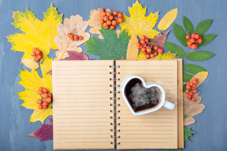 White mug in the shape of a heart with steaming coffee or tea and a blank lined craft notepad on fallen leavesの写真素材