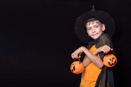 Halloween kids. Boy in wizard hat and orange t-shirt holding Halloween pumpkin-shaped buckets of sweetsの写真素材