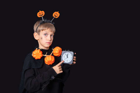 Halloween lifestyle. Boy with a spider on his cheek and a pumpkin beads holding a black alarm clockの写真素材