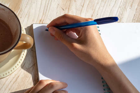 Close-up of a female hand writing down text in a ballpoint pen in a blank notebook with a cup of coffee on a tableの写真素材