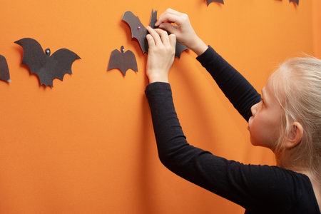 Halloween kids. Girl hanging homemade black paper-cut bat on orange background. Halloween season mood.の写真素材