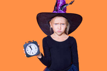 Halloween time. Sullen disgruntled girl in a fairy costume holding a black alarm clock on an orange...