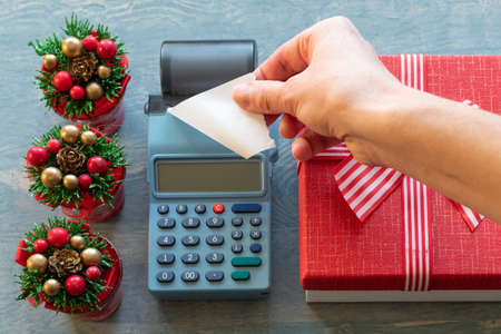 Christmas sale. Hand holding A blank white check from a cash register near a red gift box with a ribbon and small Christmas trees with cones. Buying gifts for New Year and Christmasの写真素材