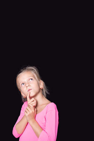 Pensive beautiful caucasian girl in a pink sweater looking up and holding her finger to her face on a black backgroundの写真素材