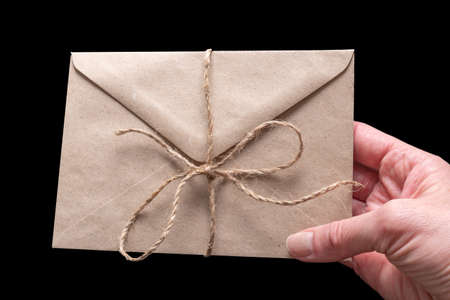 Hand holding closed brown craft envelope tied with twine isolated on black background. An envelope for your congratulationsの写真素材