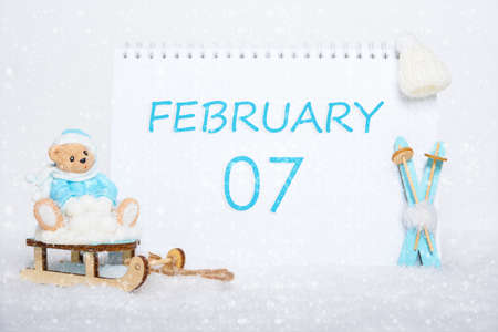 February 7th. Teddy bear sitting on a sled, blue skis and a calendar date on white snow. Day 7 of month. Winter sports concept. Winter month, day of the year concept.の写真素材