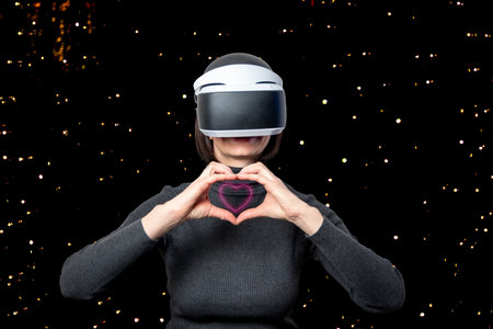 Beautiful woman using vr headset shows glowing heart shape with hands on dark background with bokeh....