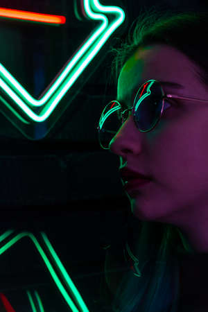 Stylish fashionable teenage girl in glasses with reflection looks up on the street with neon illumination of the cityの写真素材
