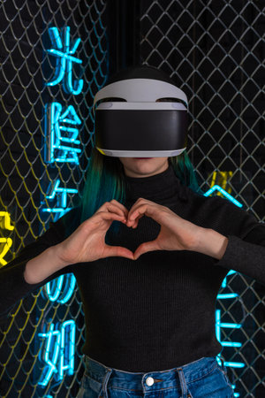 Teen girl using vr helmet shows heart gesture with hands on futuristic colorful background. Virtual...