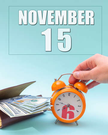 November 15th. Hand holding an orange alarm clock, a wallet with cash and a calendar date. Day 15 of month. business planning. Time is money. tax time. Autumn month, day of the year concept.の写真素材
