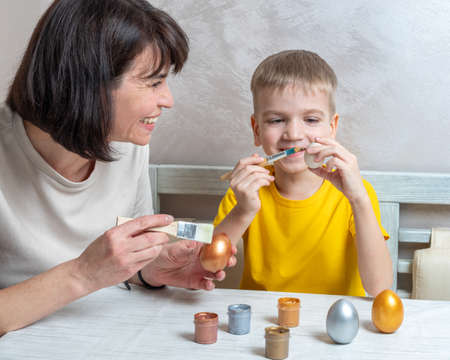 Mom and a little blond boy paint eggs with brushes for the Easter holiday in the home kitchen. Easter family holiday.の写真素材