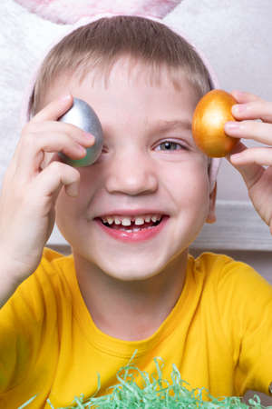 Portrait of a little blond boy covering his eyes with colorful eggs, close-up, vertical frame....