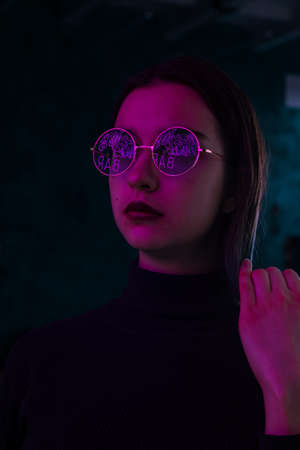 Thoughtful girl in glasses standing on the street near the evening neon lights. Stylish trendy portrait of a girlの写真素材
