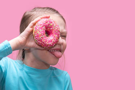 A cute funny girl in a blue t-shirt holds a bright pink donut near her eye and shows you her tongue on a pink background. Cheerful childhood concept. sweet food conceptの写真素材