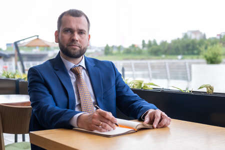 Focused middle aged 40s businessman writes notes in a personal diary, plans a working day, checks a...