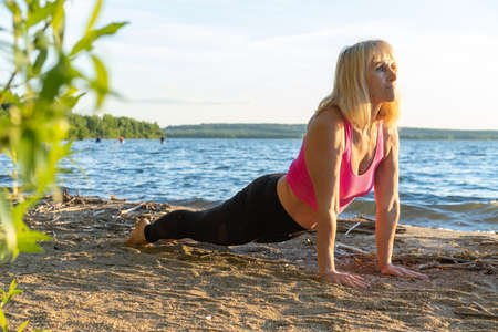 An elderly woman lifting her head up practicing yoga on the shore of a lake in summer. Meditation....