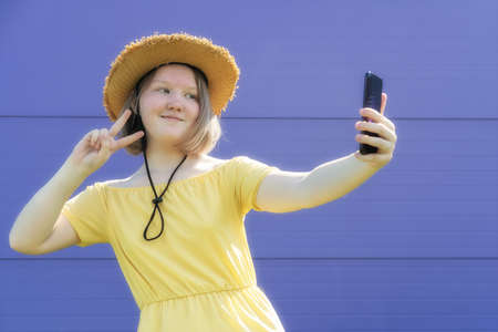 Asian teenager girl in yellow dress and hat shows greeting gesture while video chatting with friends on purple background. Trendy teenage culture. Generation Zの写真素材