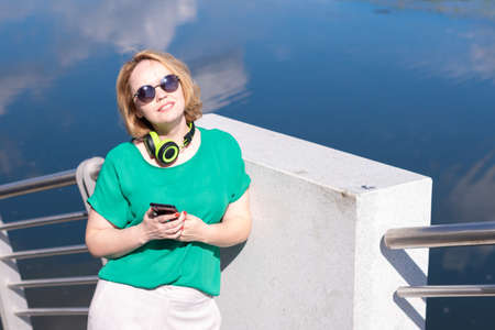 A smiling woman with headphones holding a phone in her hand listens to music standing on the riverbank. A girl with headphones and a phone on the background of the reflection of the sky with clouds.の写真素材