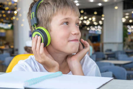 A handsome boy listens to music with headphones and writes homework in a notebook in a cafe, bokeh, close-up. Technology and music in kids life. Online remote school lesson.の写真素材