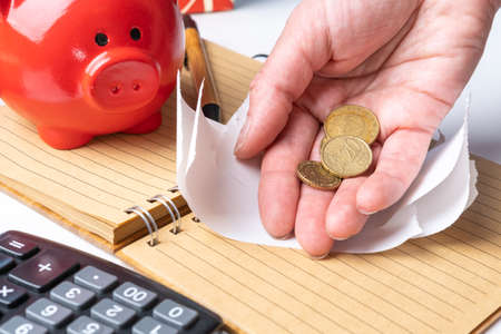 An elderly womans hand holding small coins pulled out of a piggy bank, an empty check, a calculator,...