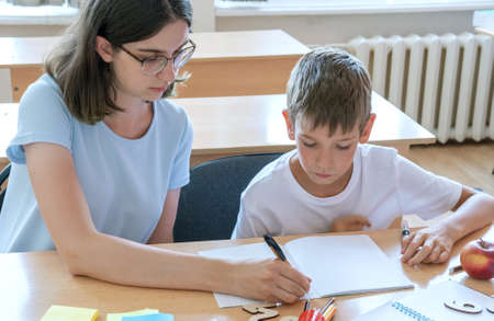 A boy and a teacher are sitting at a table with notebooks and pens. A mother helps her son write in a notebook. The babysitter helps to complete school assignments. School and preschool education.の写真素材