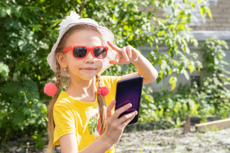 Portrait of little girl child taking a selfie by smartphone in summer park. A cheerful little girl in a yellow dress, white hat and sunglasses takes a photo on her phone and shows a V gesture.の写真素材