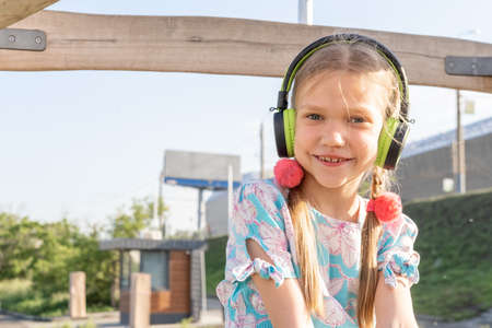 Portrait of a beautiful smiling little girl in big green headphones and a blue dress listening to her favorite music street illuminated by sunset light. Happy pigtailed girl enjoys music.の写真素材