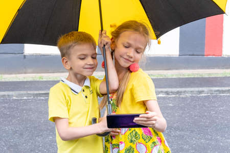 Portrait of two smiling children, a boy and a girl holding a phone, a smartphone standing under an umbrella in a park in summer. Children play an online game, taking selfiesの写真素材