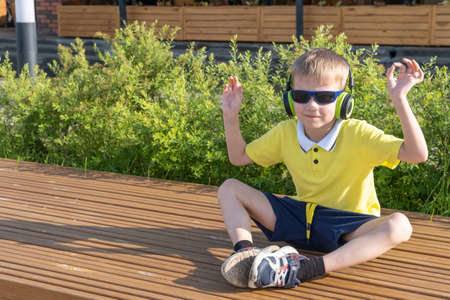A stylish boy in sunglasses with headphones listens to his favorite music while sitting on a bench in a city park. A happy smiling little boy enjoys music with his hands up.の写真素材