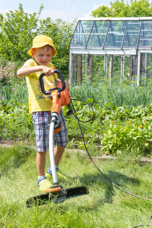 Portrait of a boy with an electric lawn mower moving the lawn. Beauty boy pruning and landscaping a...
