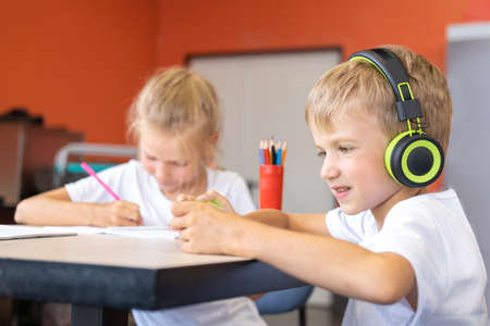 A little boy listening to music with headphones sitting at a table next to a girl student writing a text in a notebook. Online training. Back to school conceptの写真素材