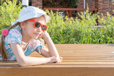 Children and summer. A happy little girl in sunglasses and a hat is laughing and smiling at the camera, lying on a bench in the city center. A cute little girl is sitting on a wooden pier.の写真素材