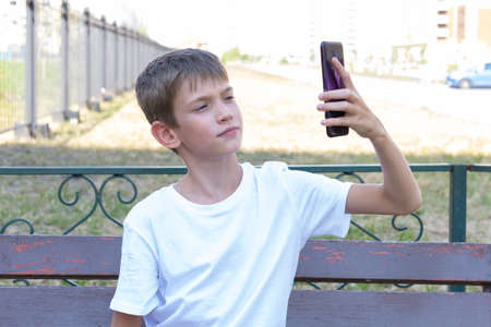 A cute boy student uses the phone, takes selfies, communicates by video call with friends, sitting on a bench on the street. online education. Back to school concept.の写真素材