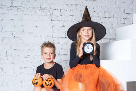Halloween time. Children in creepy witch costumes and hats are holding a black alarm clock and a...