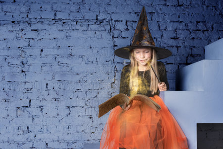 Kids Halloween. A beautiful sweet girl in a witch costume, wearing a hat, holding a book with spells and a magic wand, conjures sitting on the steps. The little sorceress sorceress is conjuringの写真素材